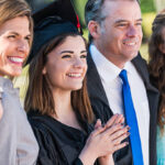 A family celebrating the graduation of their oldest daughter from high school.  They are standing together outdoors.  The teenage girl is wearing a black cap and gown with red tassel.  She is standing in the middle between her father, wearing a suit, and mother.  Her sisters are on the ends.